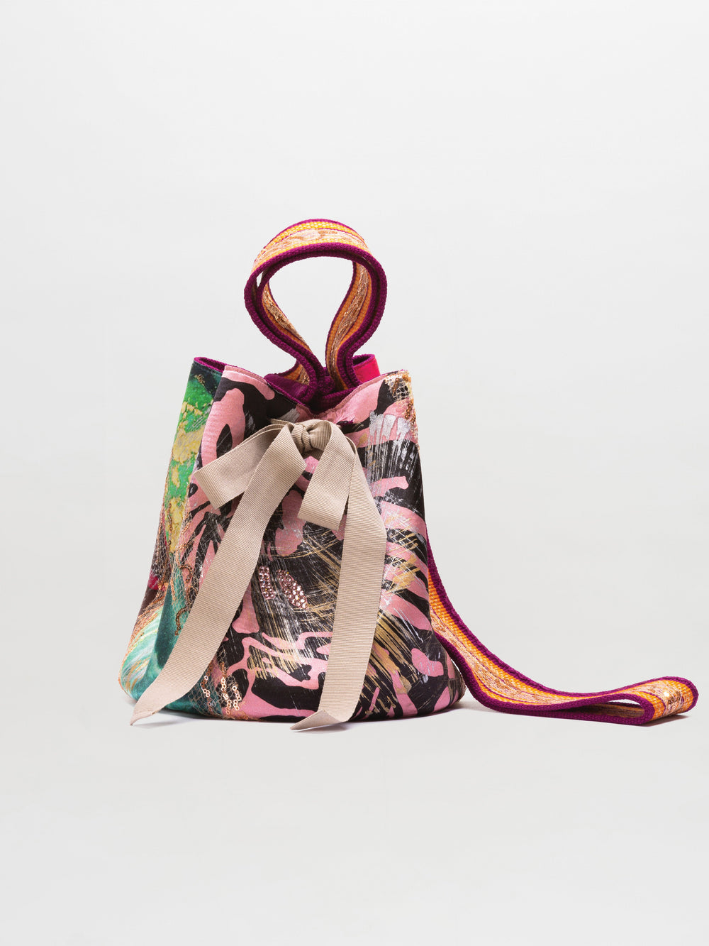 The Mochila Laaput Big is a fabric tote with multicolor abstract patterns, a beige front bow, and pink-orange trimmed handles, inspired by the vibrant style of a Wayuu mochila handbag.