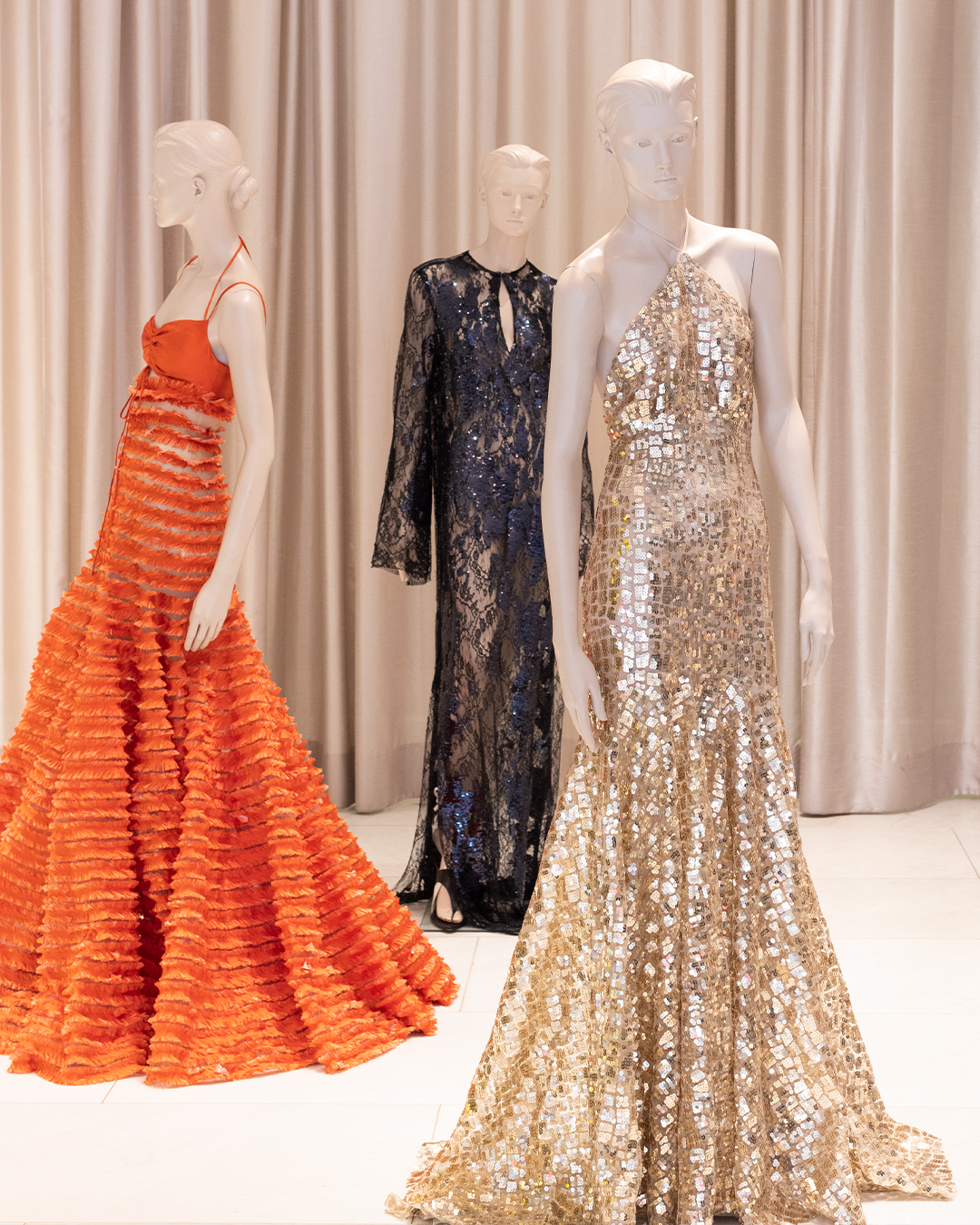 Three mannequins display evening gowns: one in a textured orange dress, one in a dark lace dress, and one in a shimmering gold sequin dress, set against a neutral curtain backdrop.