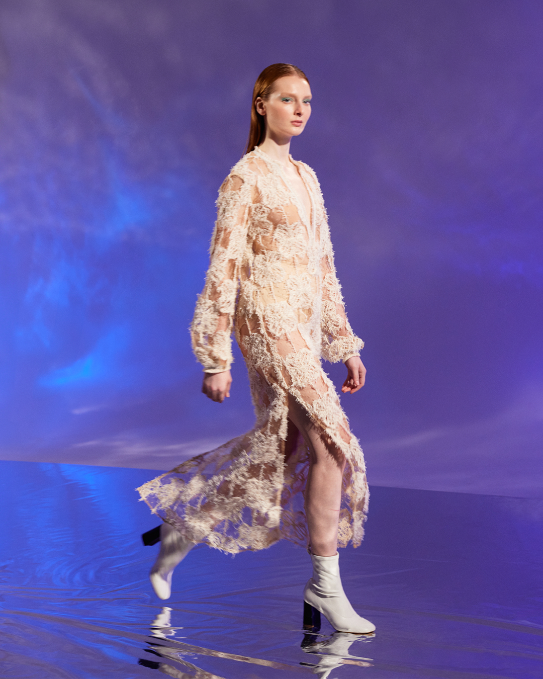 Model walks on runway in a sheer, floral lace dress and white boots against a blue background.