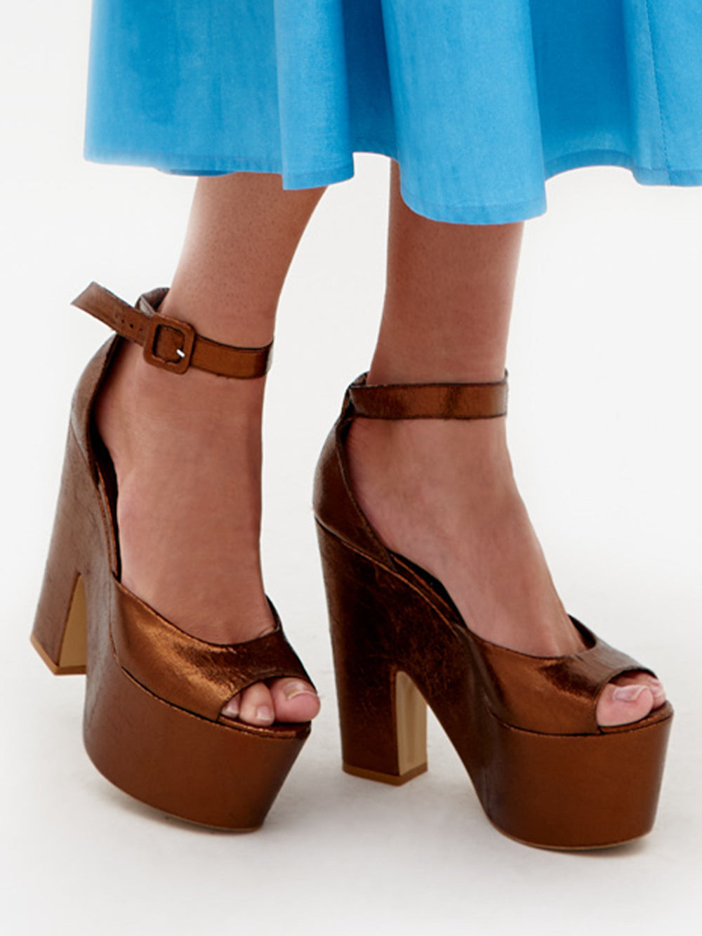 Wearing Oishi Heels Bronze platform heels with an ankle strap, a person pairs them perfectly with a light blue dress as they get ready for an elegant event, scheduled to ship by February 15th, 2025.
