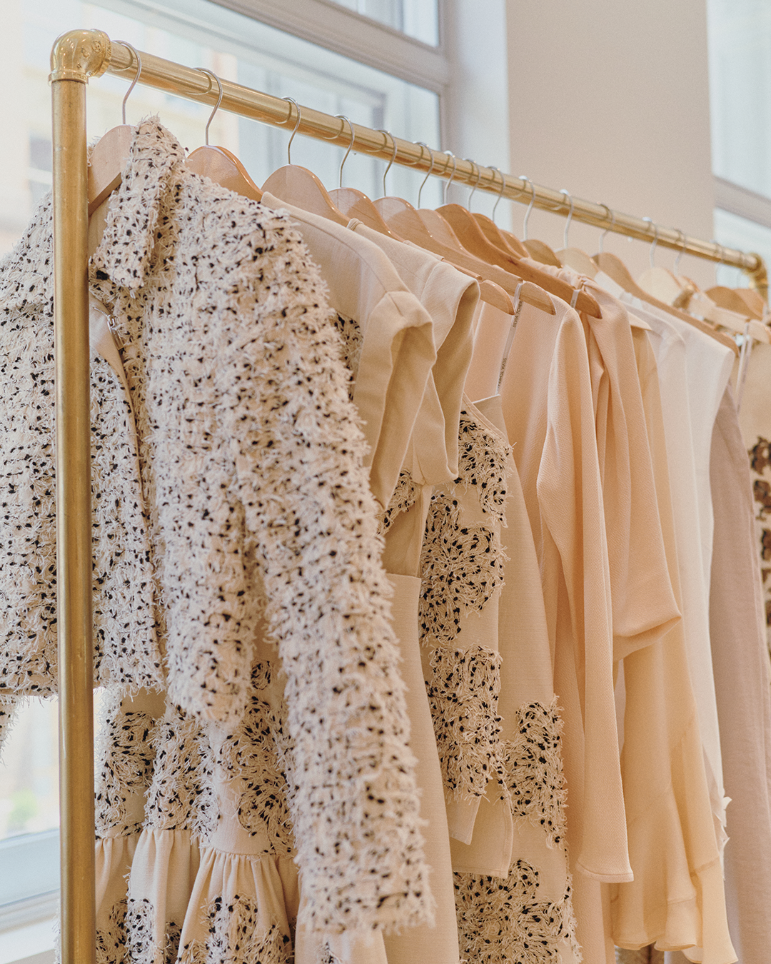 A collection of patterned and solid beige garments hangs on a gold clothing rack in a bright room.