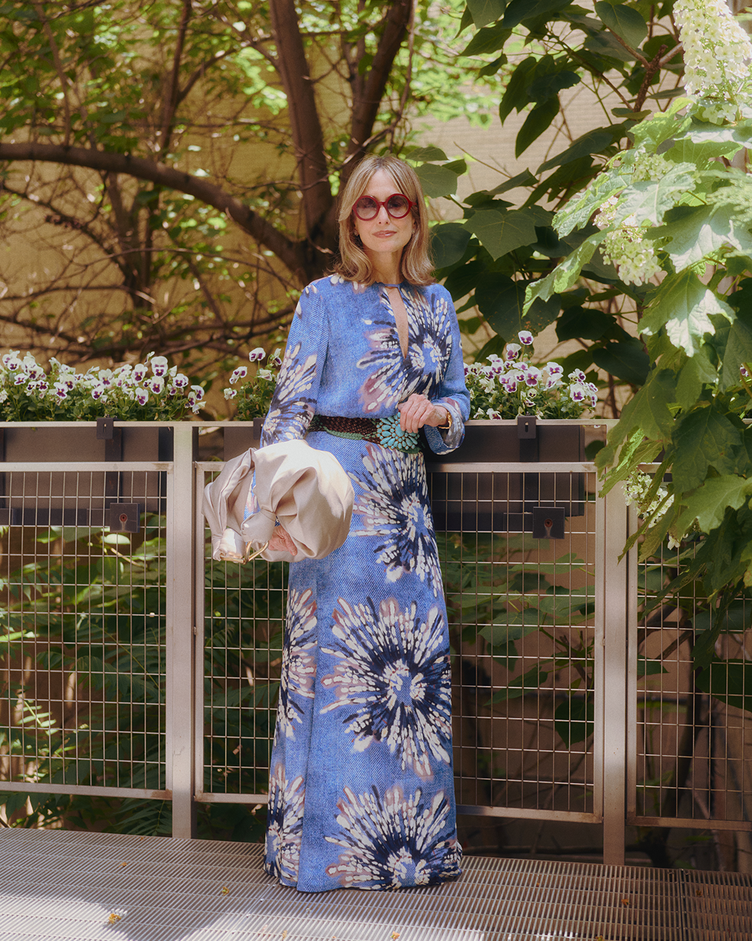 A person wearing sunglasses and a long blue patterned dress stands on a balcony surrounded by greenery, holding a beige jacket.