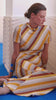 A woman in a colorful, striped dress sits thoughtfully against a textured wall. Her dress features shades of yellow, orange, and brown, complementing her calm pose.
