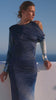 Women in a blue, patterned dress gazes into the distance against a clear blue sky and sea. The mood is serene and contemplative.