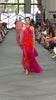 A person with blonde hair in a low ponytail walking the runway wearing the Jesse Dress—a backless, floor-length maxi with a halter neckline and a pink and red abstract pattern, made from luxurious Italian fabric—standing against a white background.