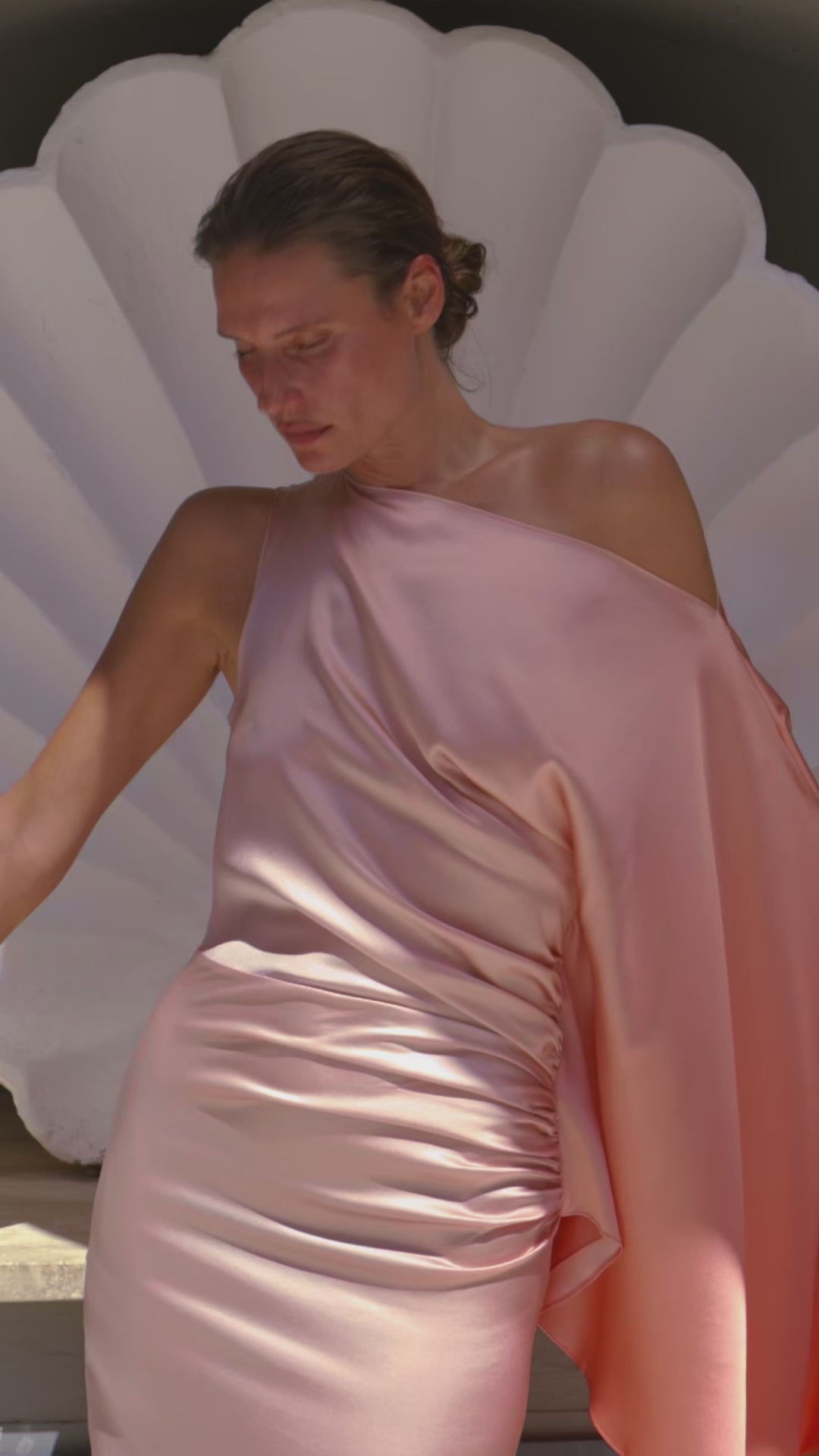 A woman in a flowing, one-shoulder pink satin dress stands pensively against a white shell backdrop. The scene exudes elegance and tranquility.