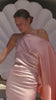 A woman in a flowing, one-shoulder pink satin dress stands pensively against a white shell backdrop. The scene exudes elegance and tranquility.
