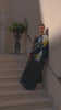 A women in a one-shouldered, abstract-patterned dress leans against a wall. They wear chunky jewelry and gaze thoughtfully, conveying a serene yet intense mood.