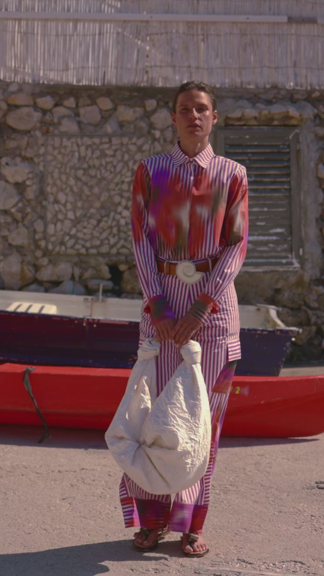 A person stands in front of a stone wall and red boat, wearing a colorful, striped outfit and holding the Sofia Bag off-white. The mood is calm and stylish.