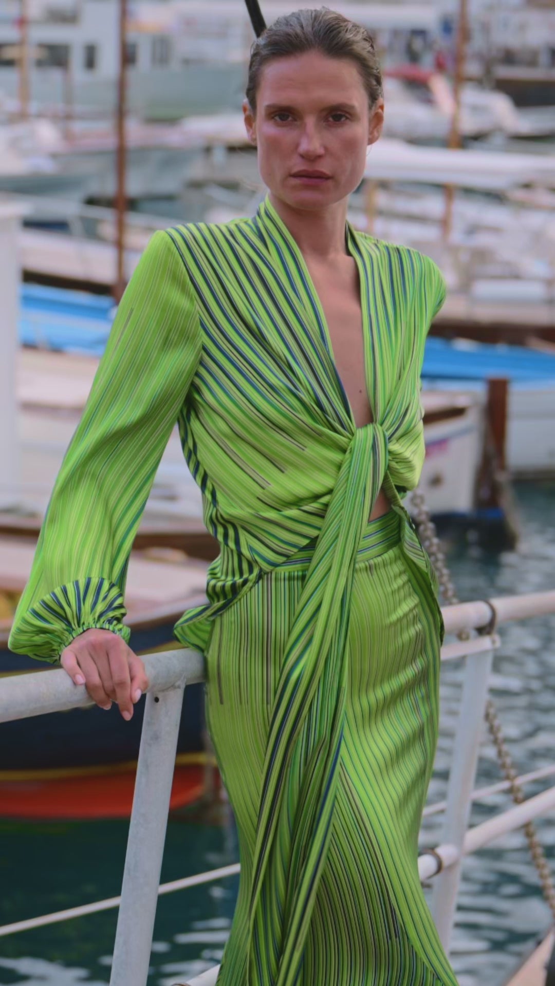 A women stands on a dock wearing a green, striped outfit. The background features blurred boats and water, creating a calm, nautical atmosphere.