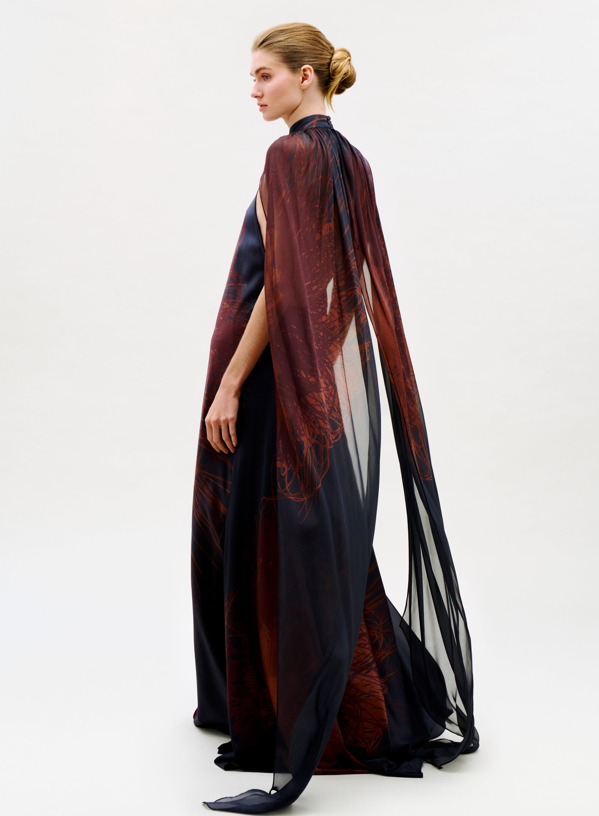 A woman with blonde hair in a low bun models the Sabina Dress, a flowing sheer black and dark red gown with a dramatic cape, standing in profile against a plain white background.