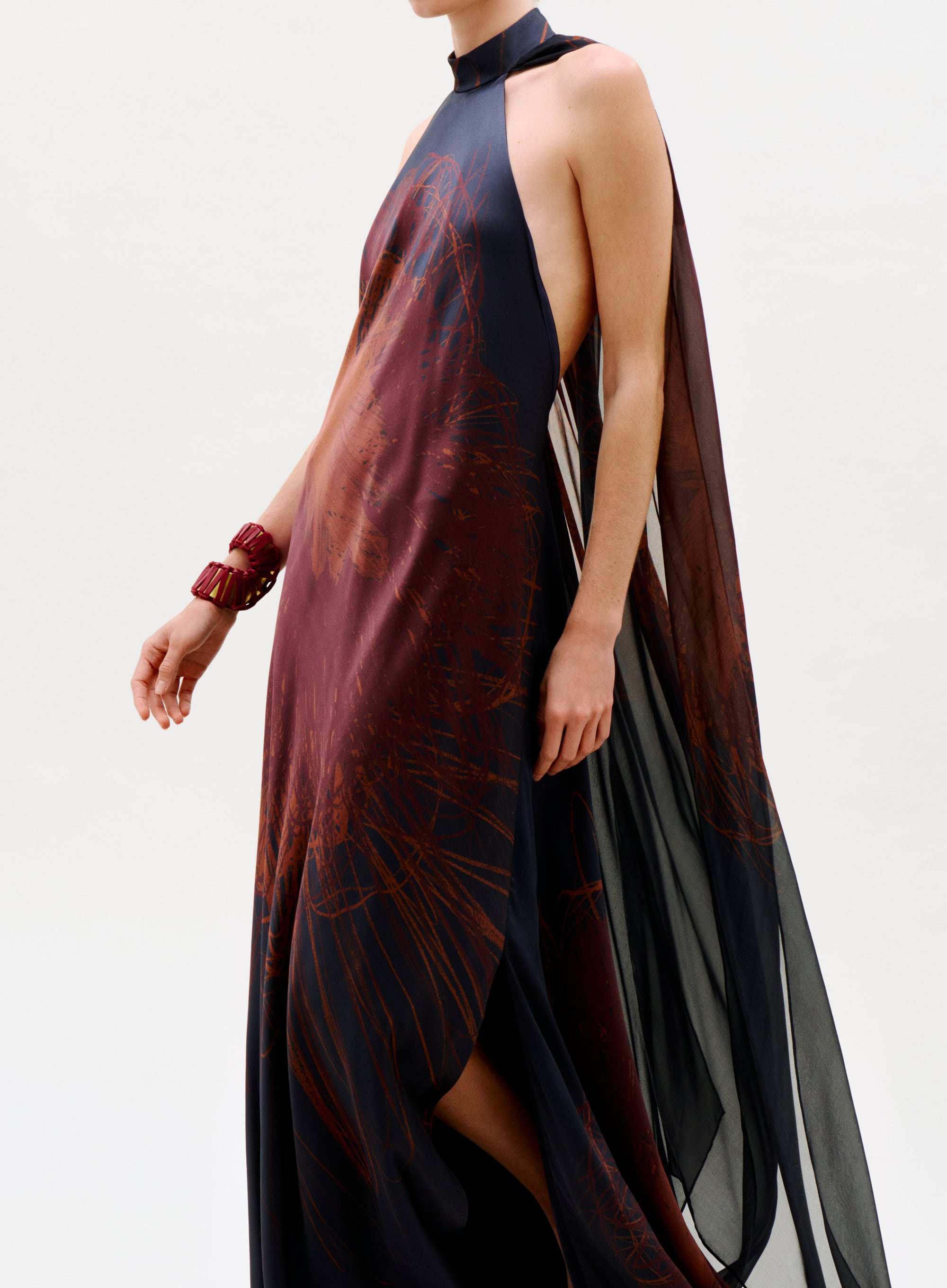 A person wearing the Sabina Dress—a sleeveless, high-neck black and deep red sheer gown with cape-like draping—paired with a red bracelet, poses against a plain light background.