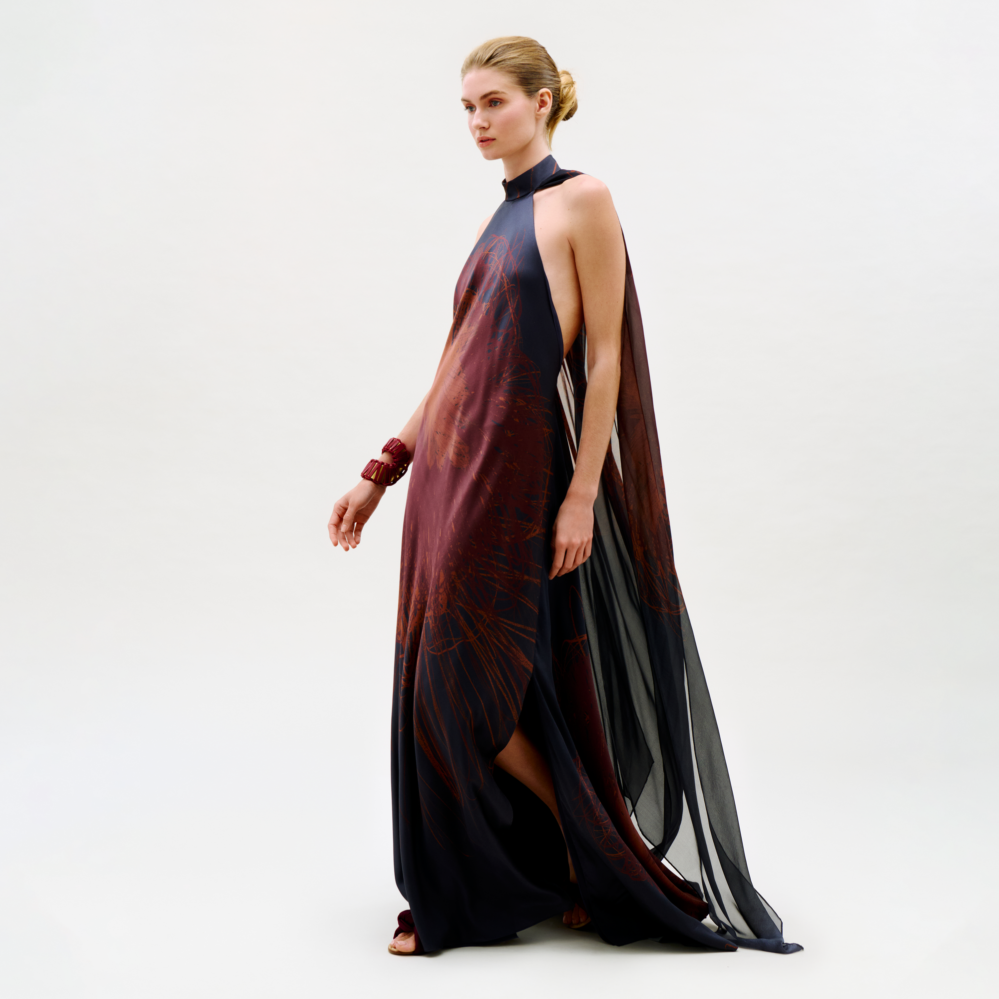 A woman poses against a neutral background in the Sabina Dress, a sleeveless, floor-length dark gown featuring side slits and sheer panels. She accessorizes with a chunky bracelet and has her hair styled in a neat updo.