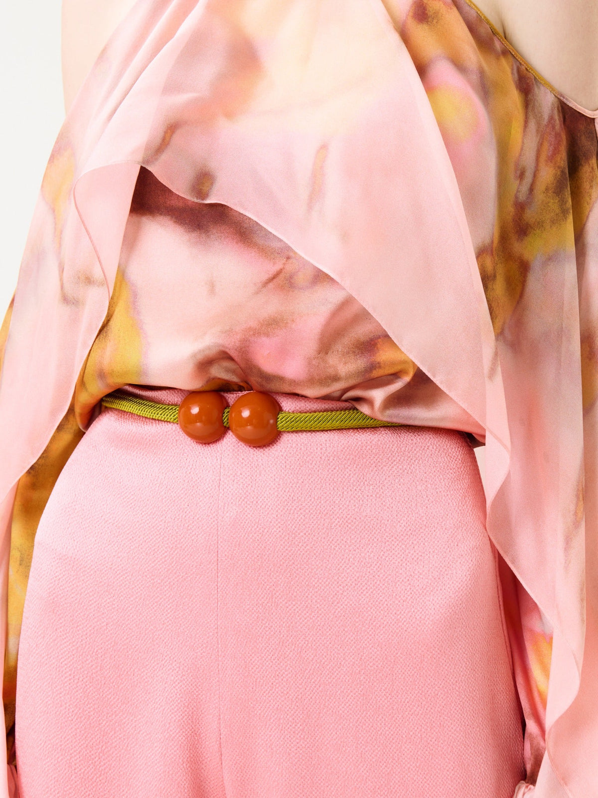 A person wearing the Shaan Blouse—a flowing, pink patterned top—paired with pink pants and a thin green belt featuring two round orange beads at the waist.