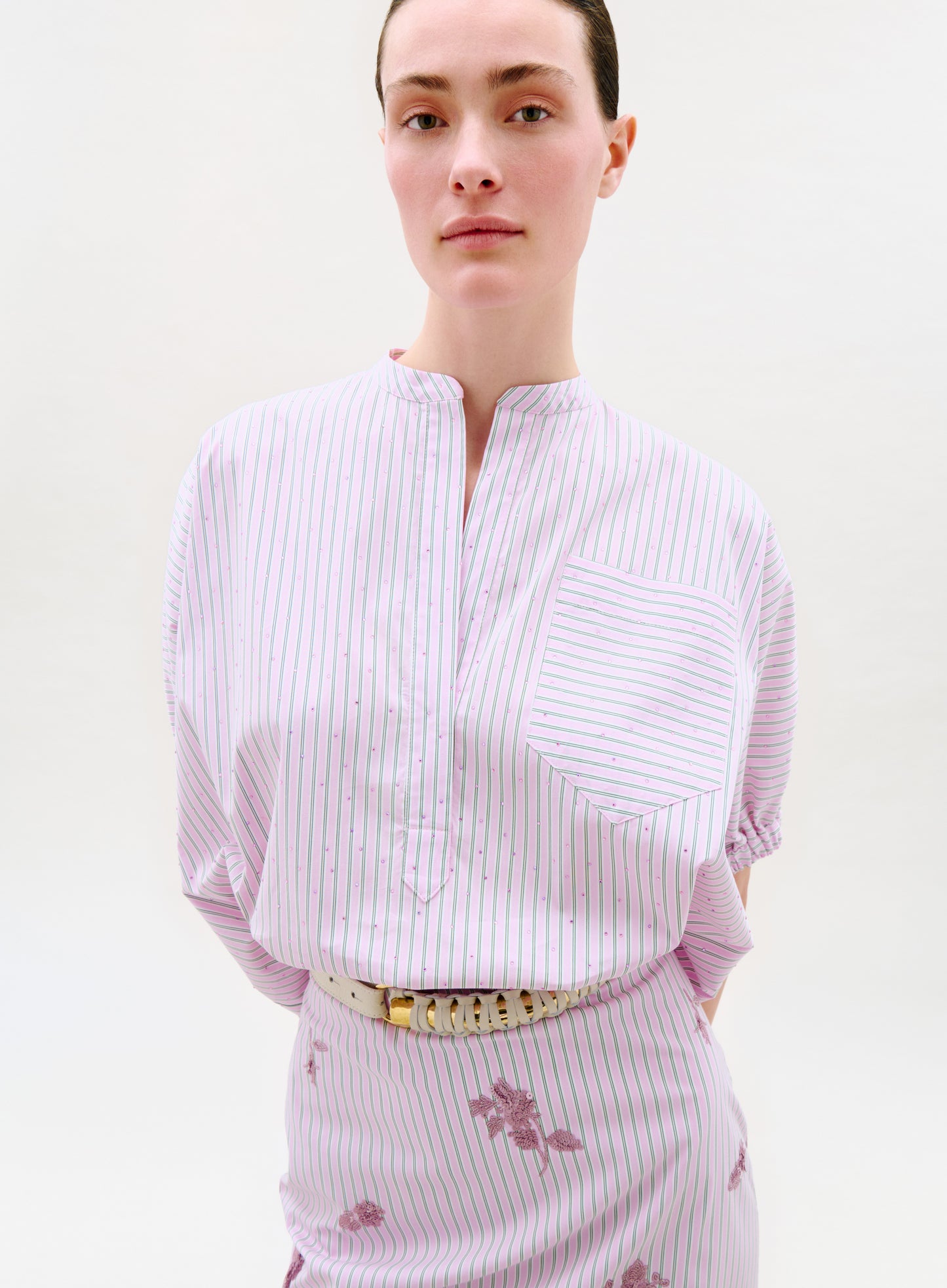 A person wears the Susane Blouse, a short-sleeve top made from Italian fabric with thin pink and green vertical stripes, a front pocket with horizontal stripes, a band collar, and a cream belt. The background is plain and light.
