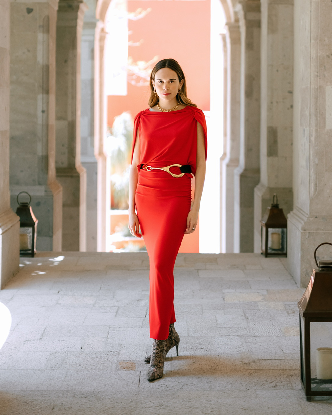 A woman in a red dress stands in a sunlit stone hallway with arches, wearing patterned boots and a belt with a large buckle. Lanterns are placed along the sides.