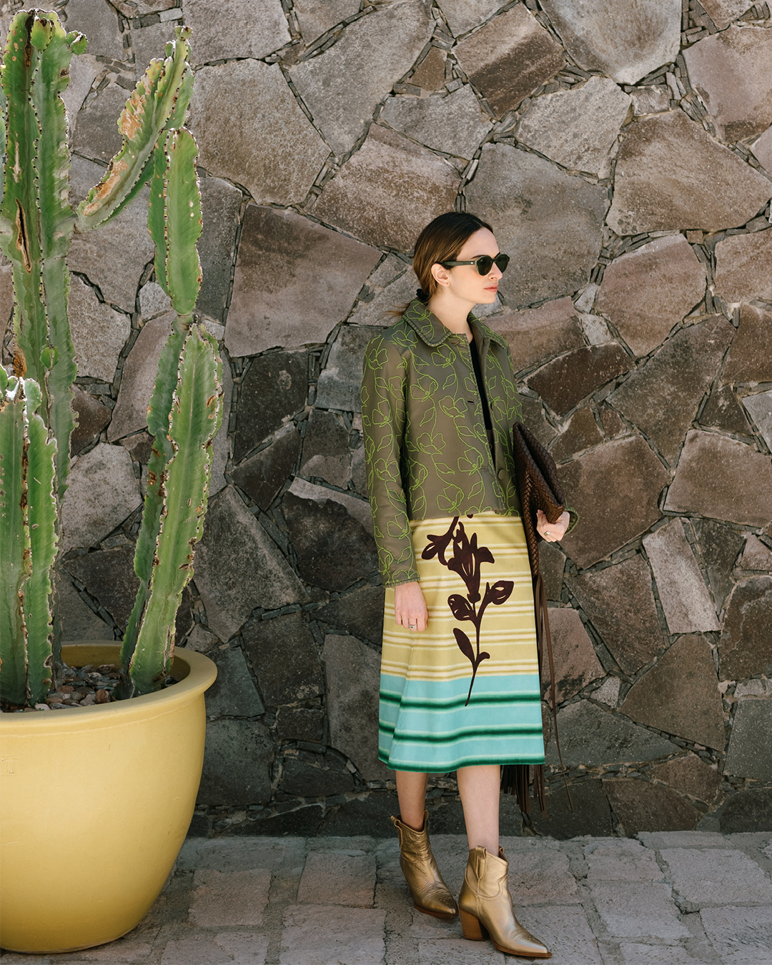 Person in a patterned green and yellow dress stands beside a potted cactus on a stone wall background.