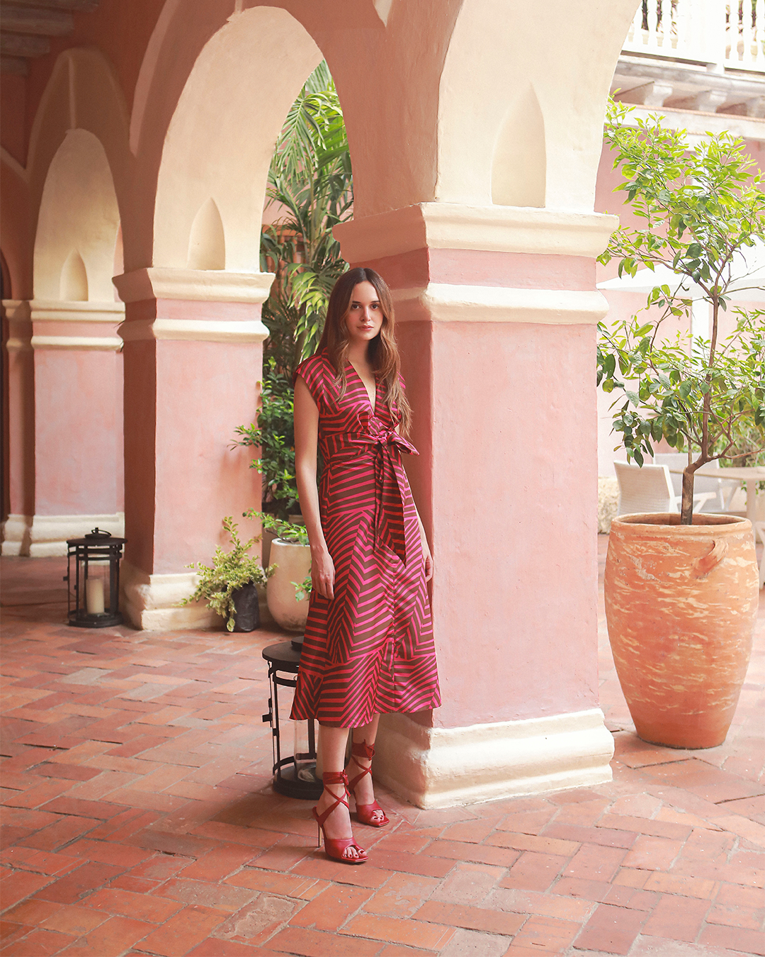 Woman in a red dress stands in a terracotta-tiled courtyard with pink arches and large potted plants.