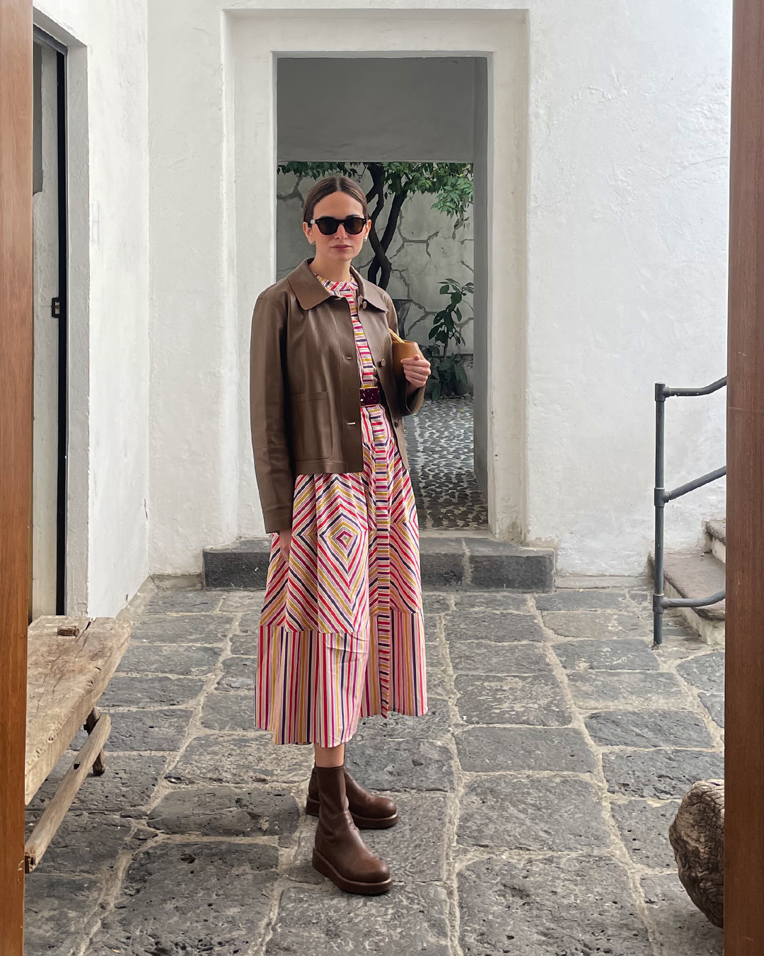 A woman in sunglasses wearing a brown jacket and patterned dress stands on a stone patio in front of a white wall.