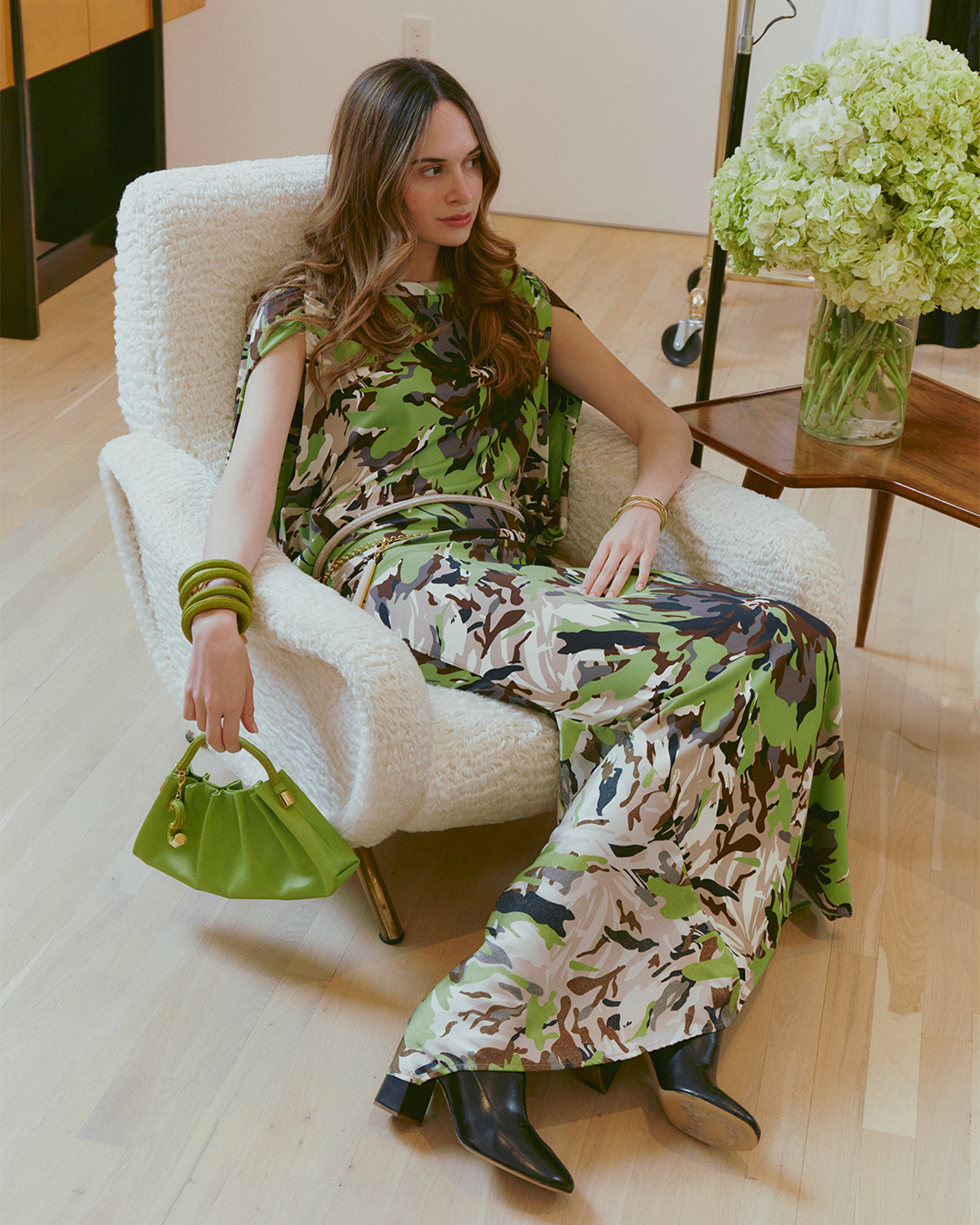 A person seated in a chair wearing a green and brown camouflage-patterned dress, holding a green handbag, with a vase of green hydrangeas on a nearby table.