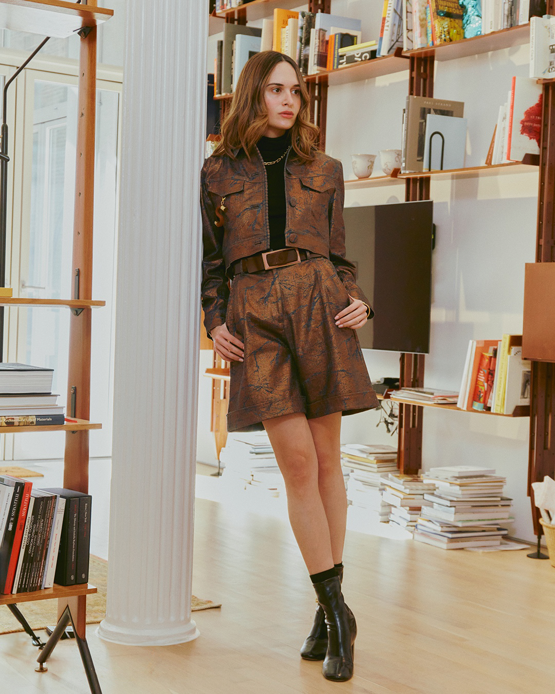 Person standing in a room with bookshelves, wearing a patterned outfit and black boots.