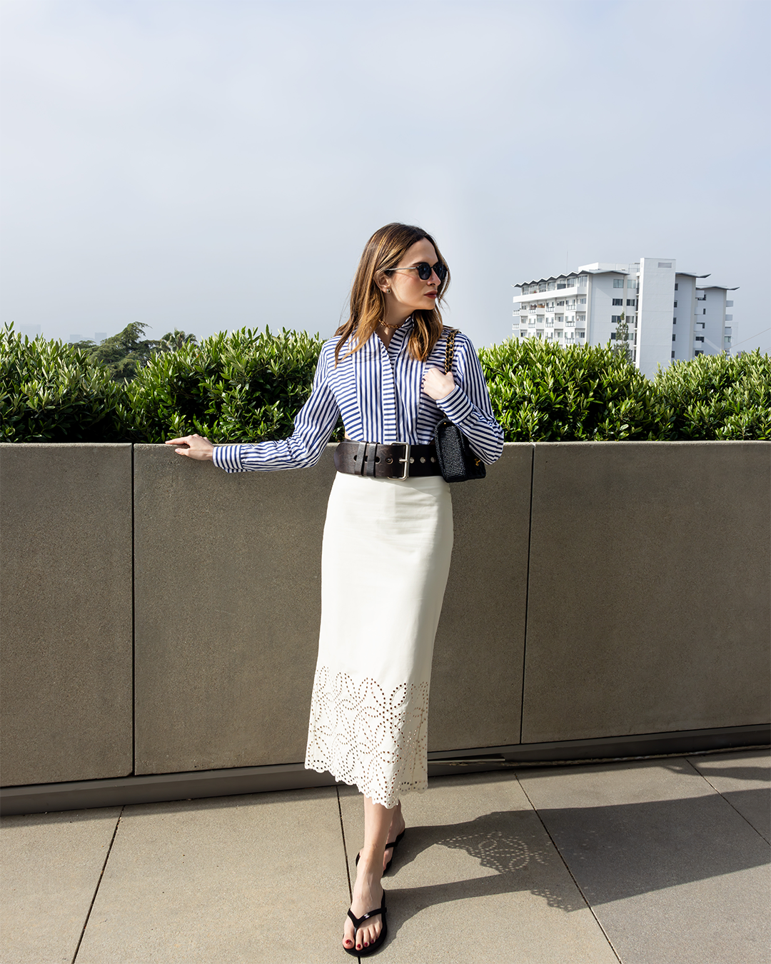 A woman stands on a terrace, wearing a striped shirt, white skirt, black belt, and sunglasses. There are green shrubs and a building in the background.