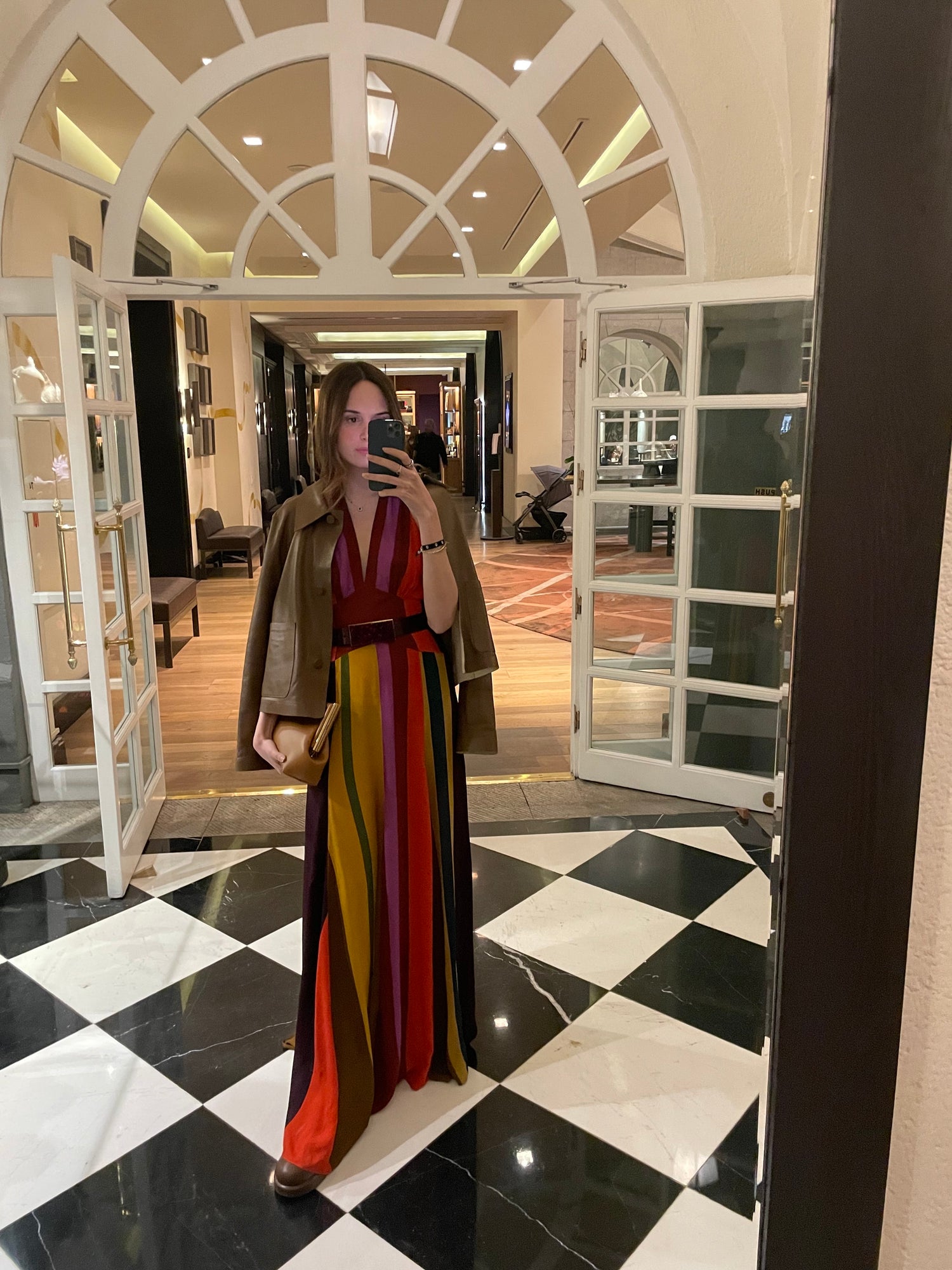 A person takes a mirror selfie in a hallway, wearing a colorful striped dress and holding a clutch. Double doors and checkered flooring are visible.