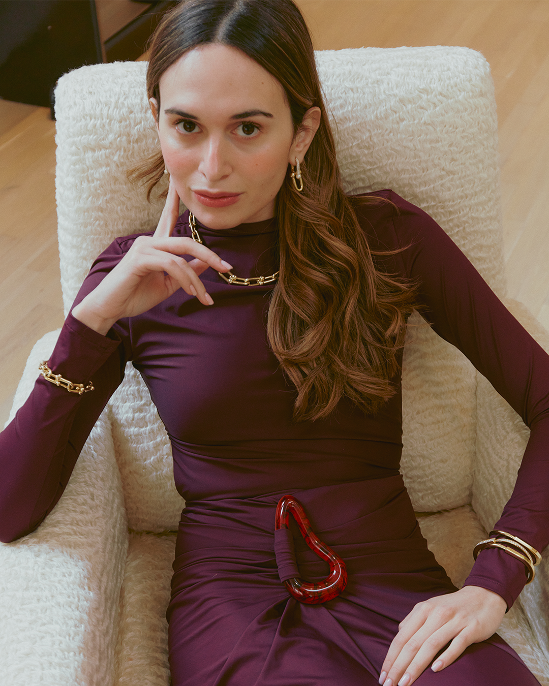 Person wearing a purple outfit with gold jewelry sits in a white textured chair, touching their face with one hand and resting the other on their lap.