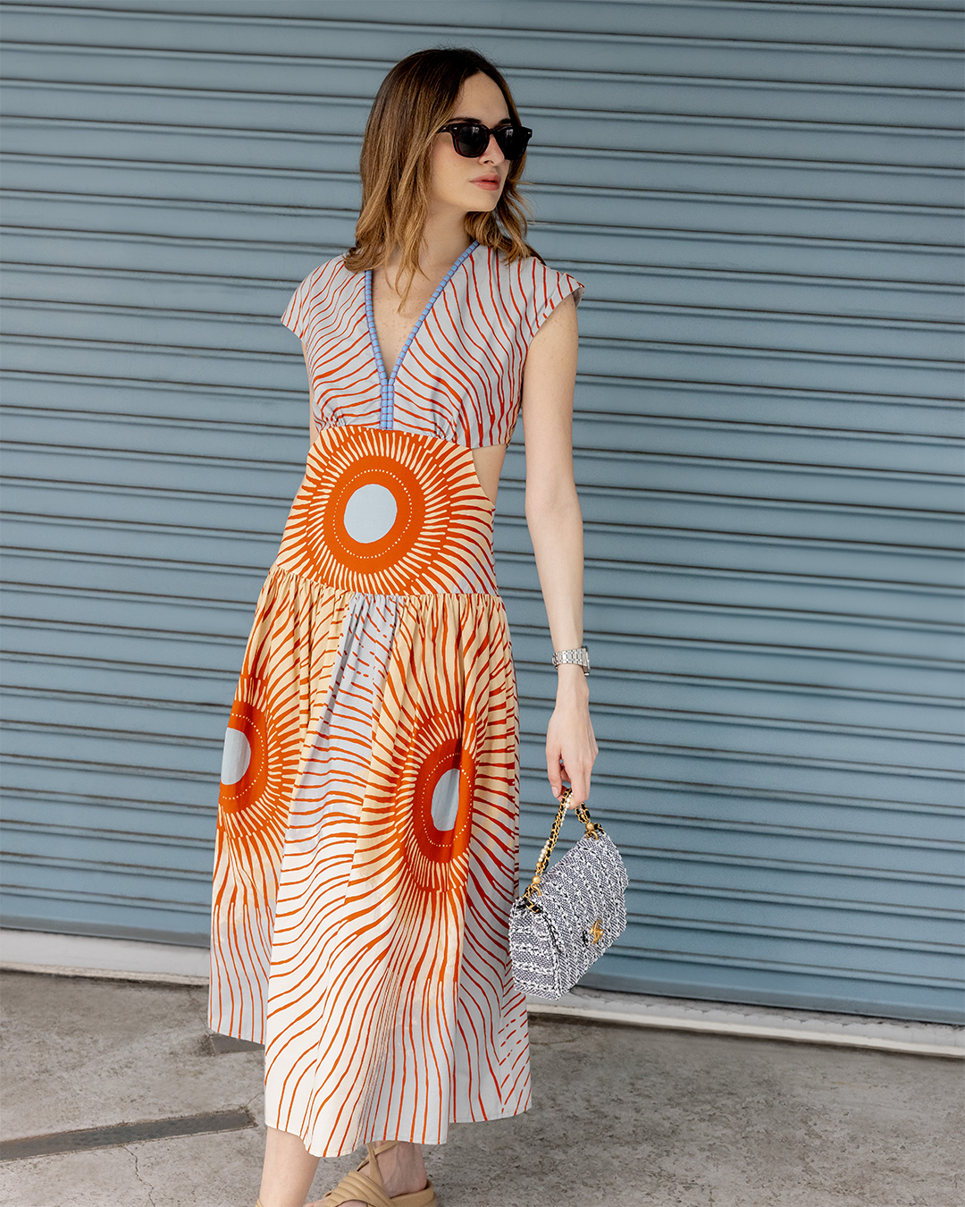 A woman wearing sunglasses and a colorful patterned dress stands in front of a closed blue shutter door, holding a small handbag.