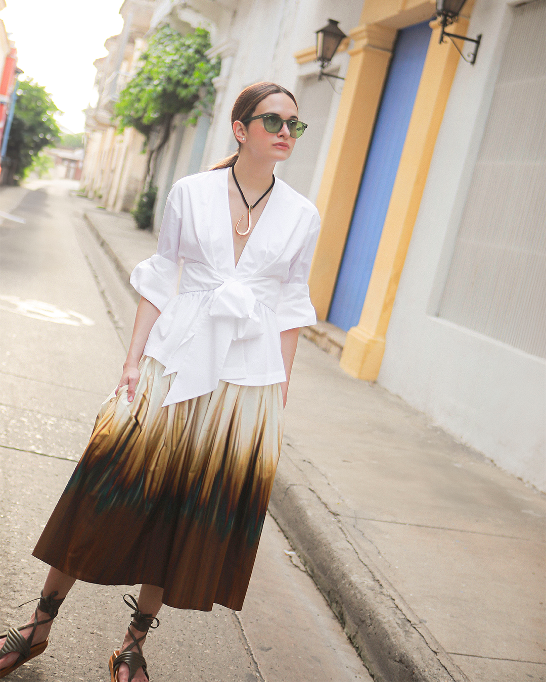 A person wearing sunglasses, a white blouse, and a long ombre skirt walks down a quiet street with colorful buildings.