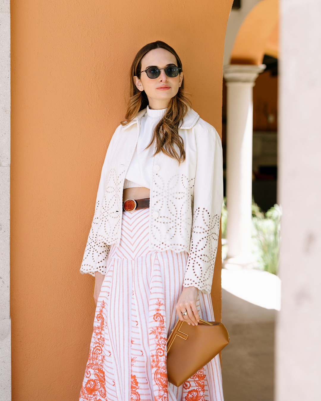 Woman with sunglasses and long hair in a white jacket and patterned skirt, holding a tan clutch, stands by an orange wall.