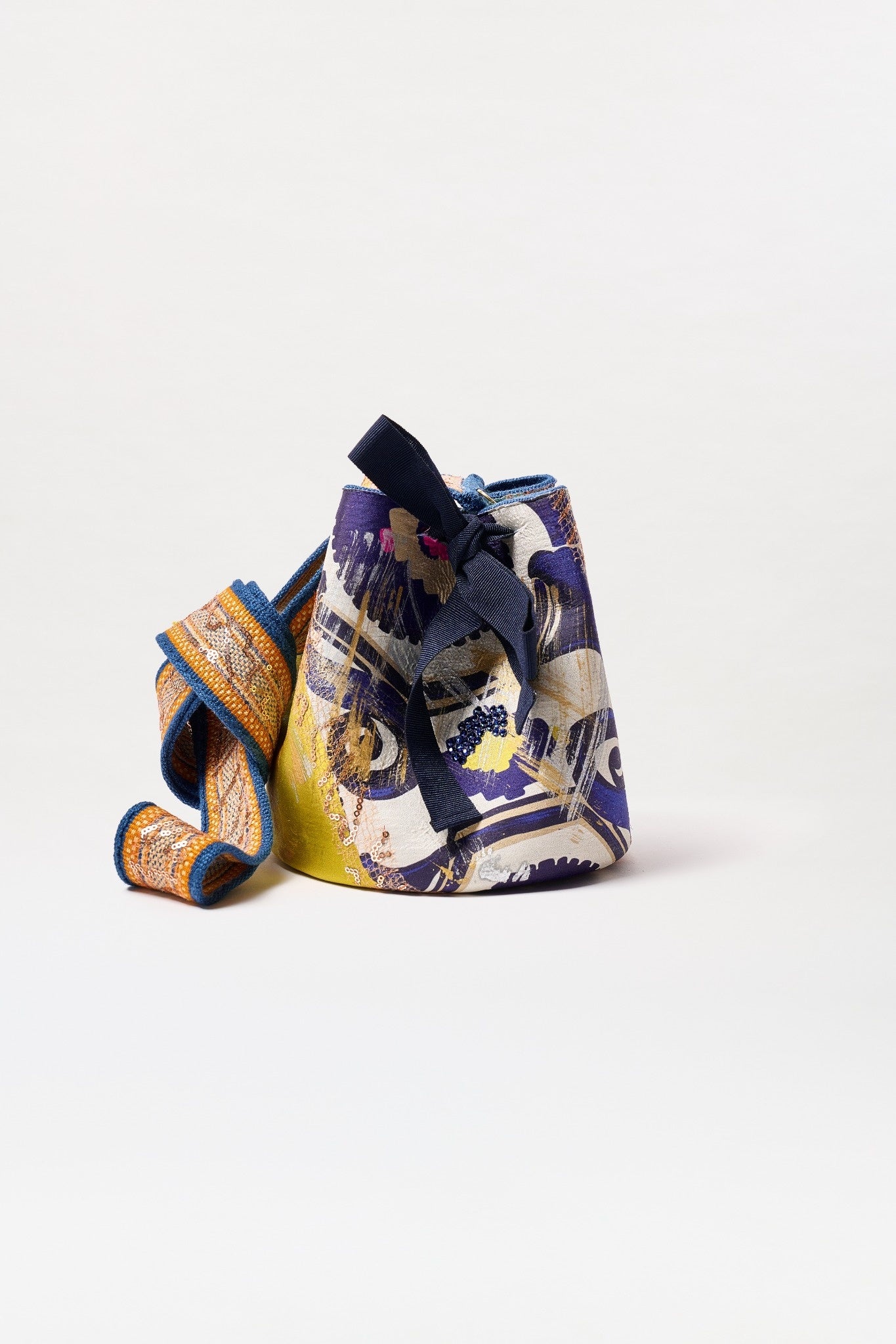 The Mochila Laaput Small is a colorful Colombian bucket bag with abstract purple, yellow, and white patterns, a thick patterned shoulder strap, black drawstring closure with bow, shown on a plain white background.