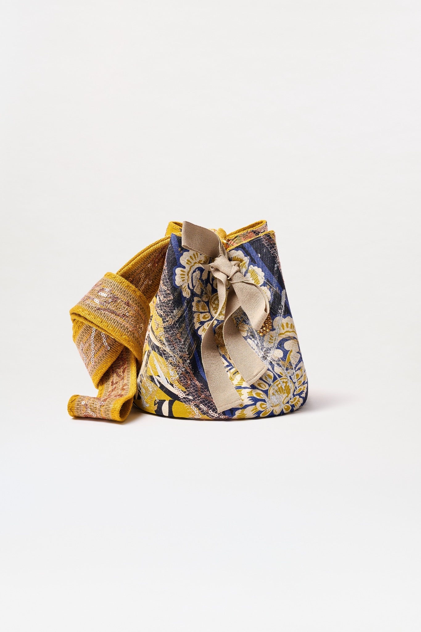 The Mochila Laaput Small is a Colombian-made bucket bag with a bold blue, yellow, and gold floral pattern. It has a wide textured yellow handle and a decorative bow tied at the top.