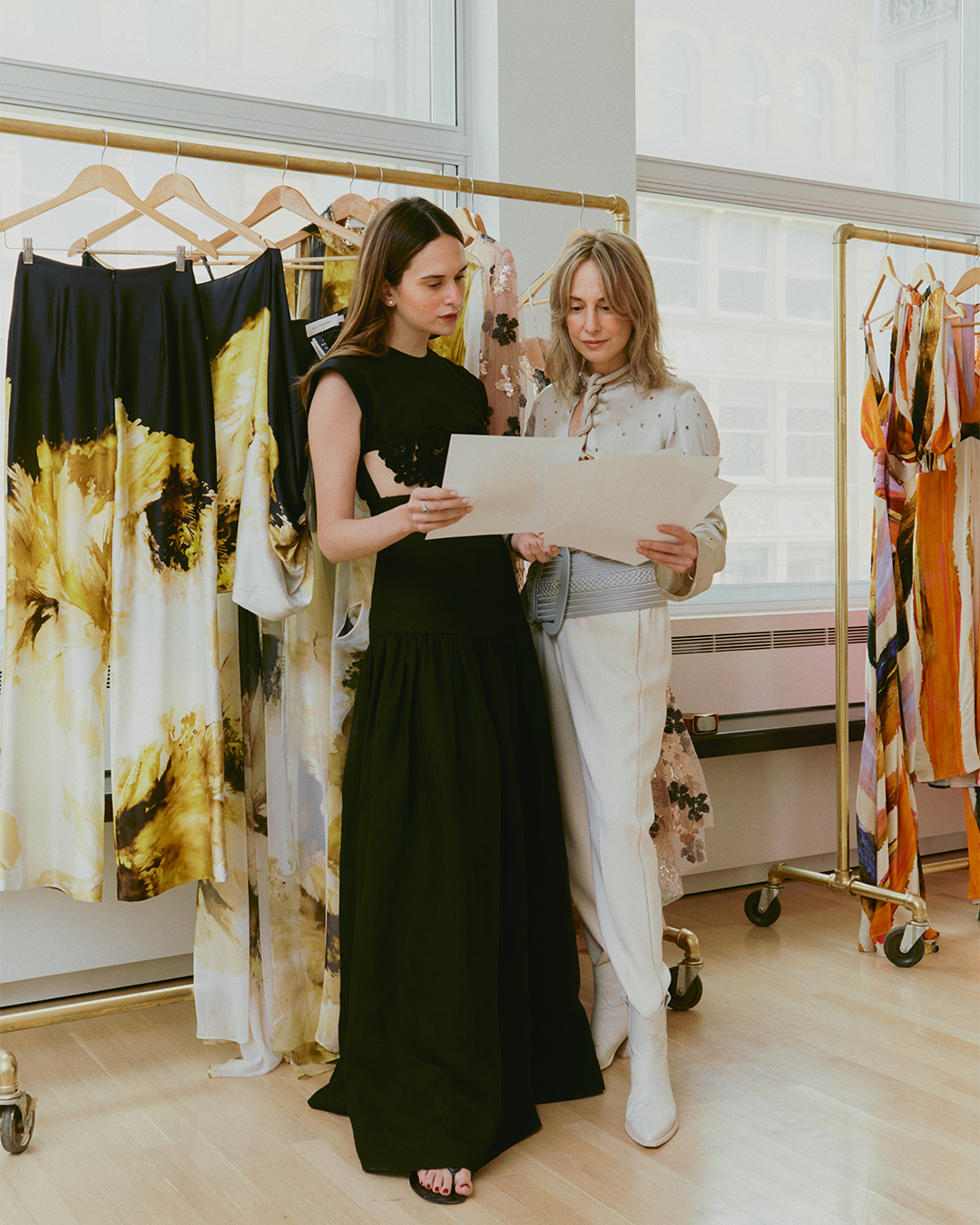 Two women stand in a room with racks of colorful clothes, reviewing papers together.