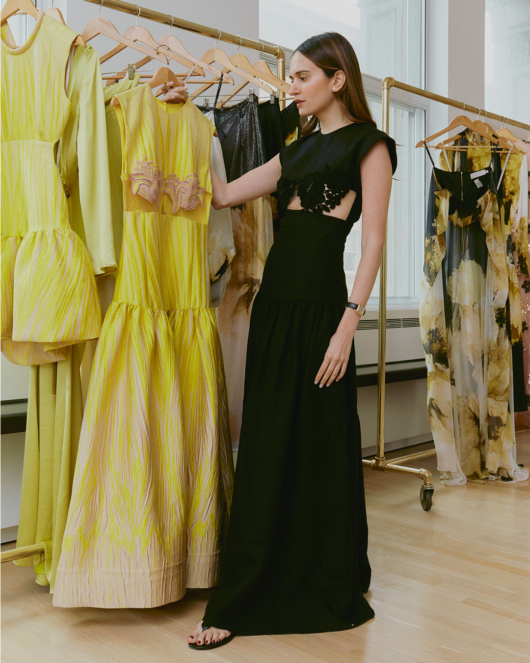 A woman in a black dress examines a yellow dress on a rack in a room with multiple garments hanging on gold clothing racks.