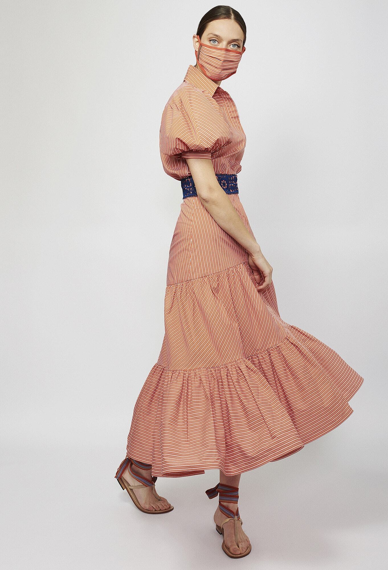 A woman in a striped red dress and matching mask poses against a white background. She wears sandals and a blue belt.