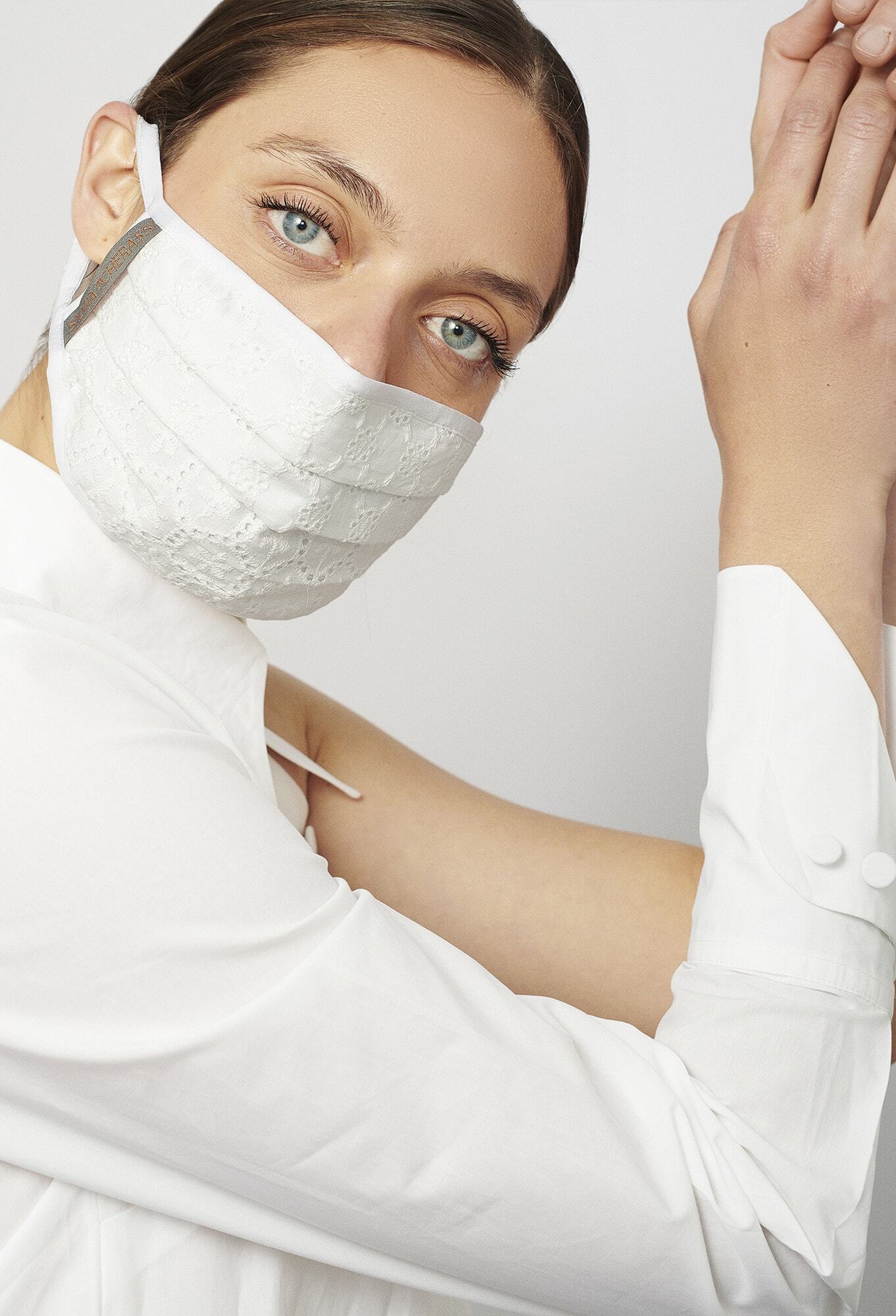 Person in a white shirt wearing a white face mask, with eyes looking directly at the camera and hands raised.