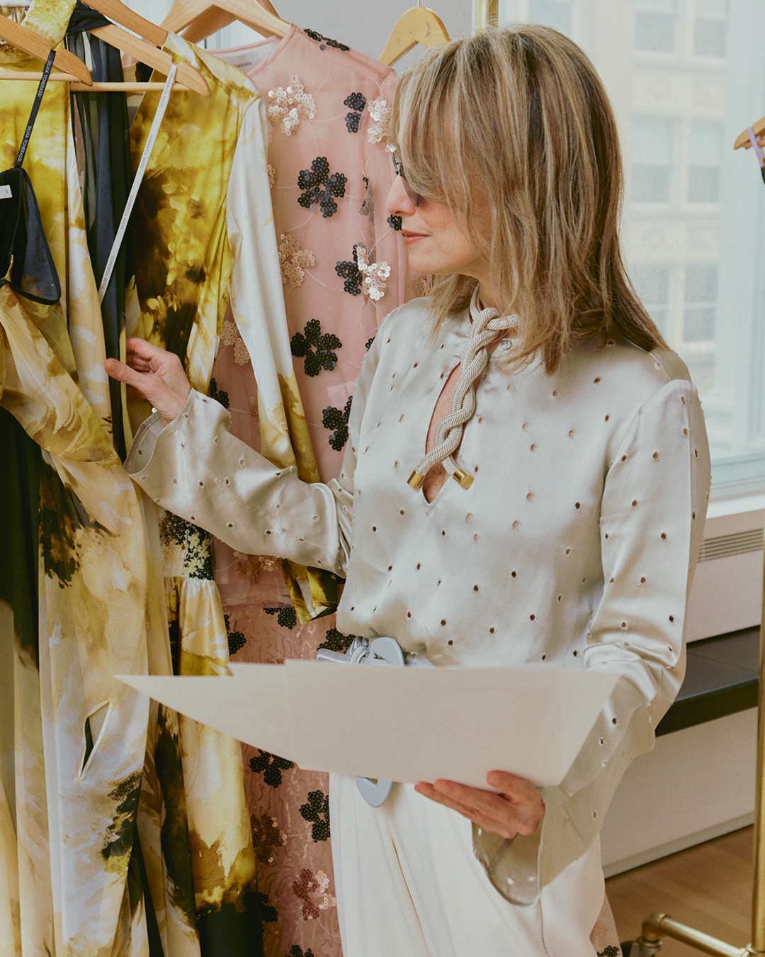 A woman in a light blouse examines garments on a rack while holding papers.