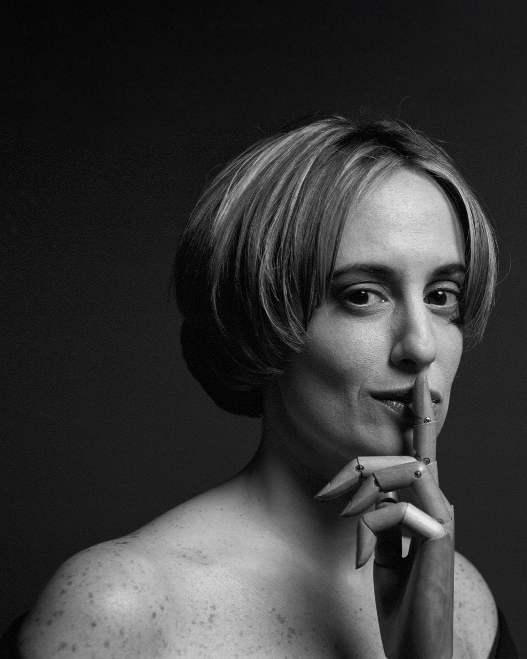 A woman with short hair holds a prosthetic hand to her lips, suggesting silence, against a dark background.