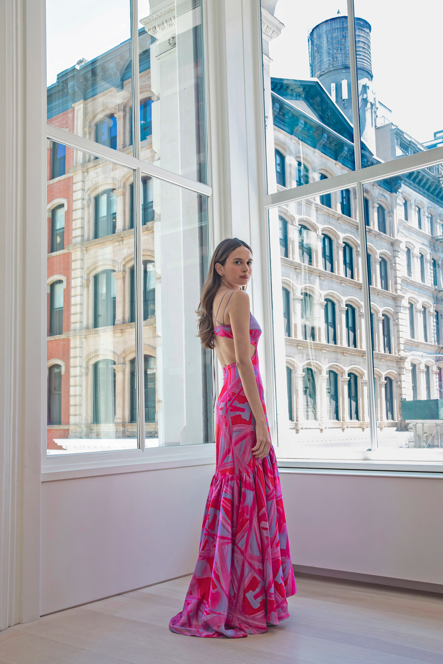 woman standing in front of a large window, wearing a vibrant pink and red dress with abstract patterns. She is positioned with her back slightly turned toward the camera, looking over her shoulder. The background features a view of city buildings with large windows. 