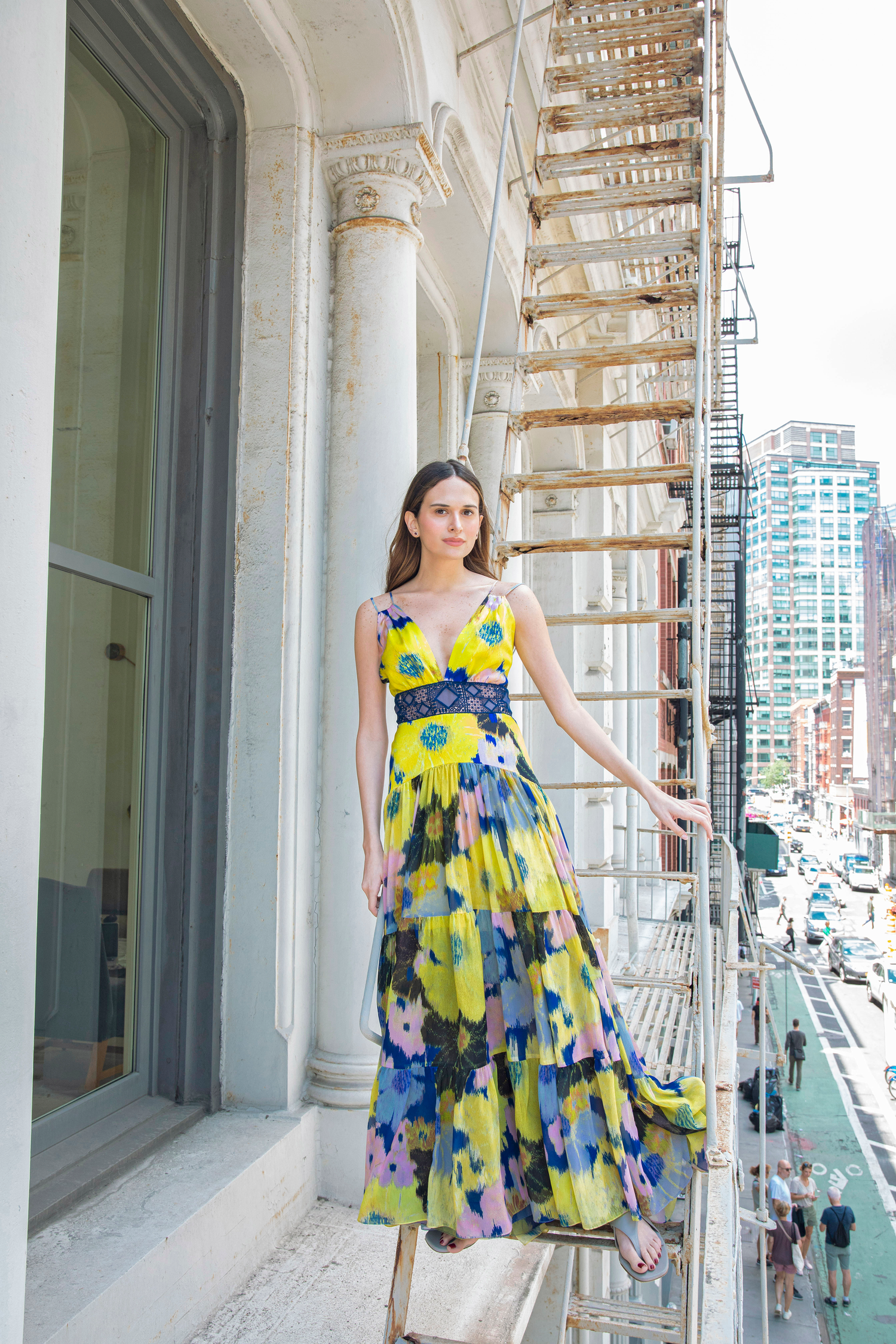 a woman standing on an outdoor fire escape, wearing a vibrant, multicolored floral maxi dress. The dress features large yellow, blue, and pink flowers and flows in layers. She is facing forward with her arms at her sides, and the background includes city buildings and a street scene with cars and pedestrians. 