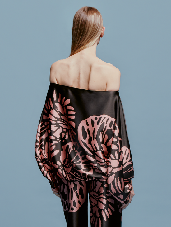 A woman is wearing the Bellagio Blouse Black Pink Filigrana, holding a black handbag, and standing against a plain light blue background, capturing the essence of late summer style just in time for September 2nd.