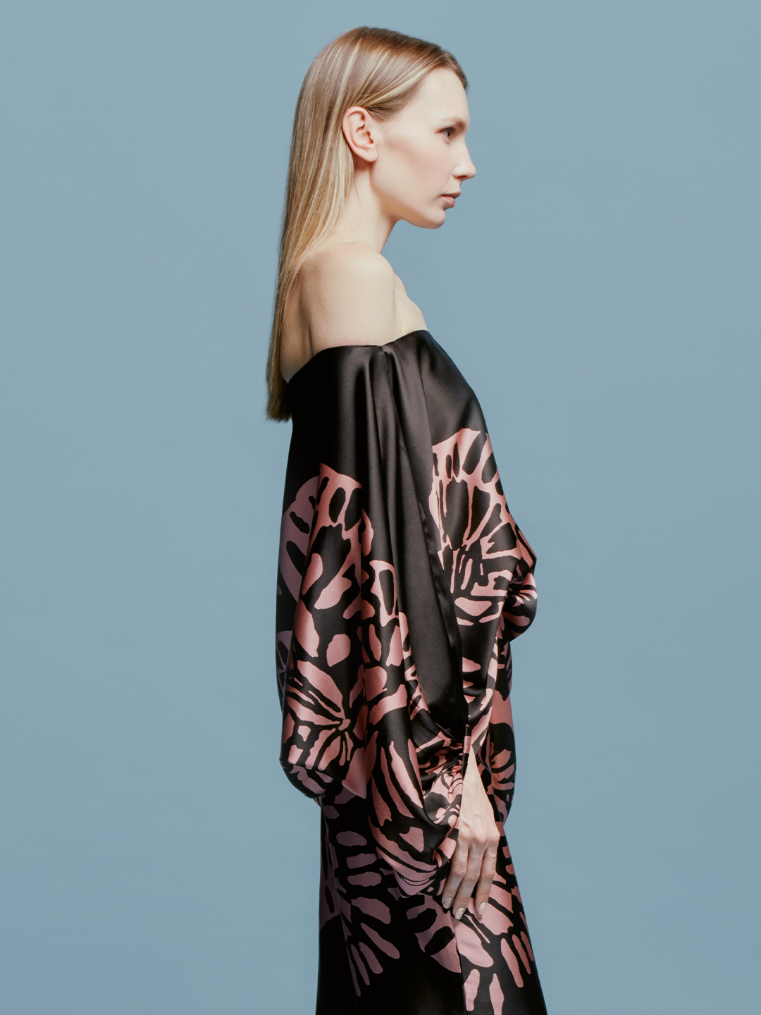 A woman is wearing the Bellagio Blouse Black Pink Filigrana, holding a black handbag, and standing against a plain light blue background, capturing the essence of late summer style just in time for September 2nd.