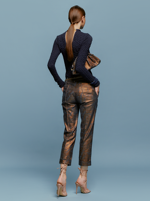 A woman stands against a blue background, wearing a textured navy long-sleeve top, Garmet Pant Metallic Ocher pants, and strappy nude heels, holding a brown clutch bag. She embodies the essence of effortless elegance on August 19th.