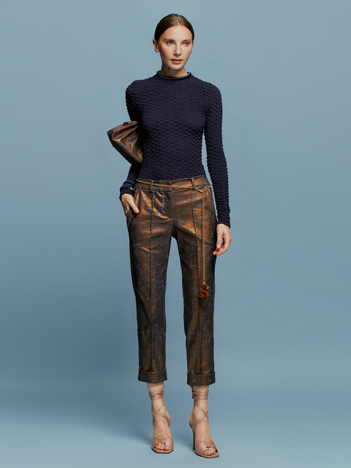 A woman stands against a blue background, wearing a textured navy long-sleeve top, Garmet Pant Metallic Ocher pants, and strappy nude heels, holding a brown clutch bag. She embodies the essence of effortless elegance on August 19th.