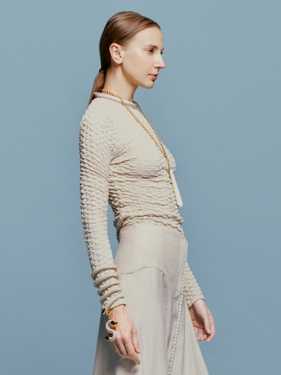 A woman with tied-back hair wears the Jari Top Cream, a long gold necklace, and several bracelets, standing against a light blue background reminiscent of the sky on August 19th.