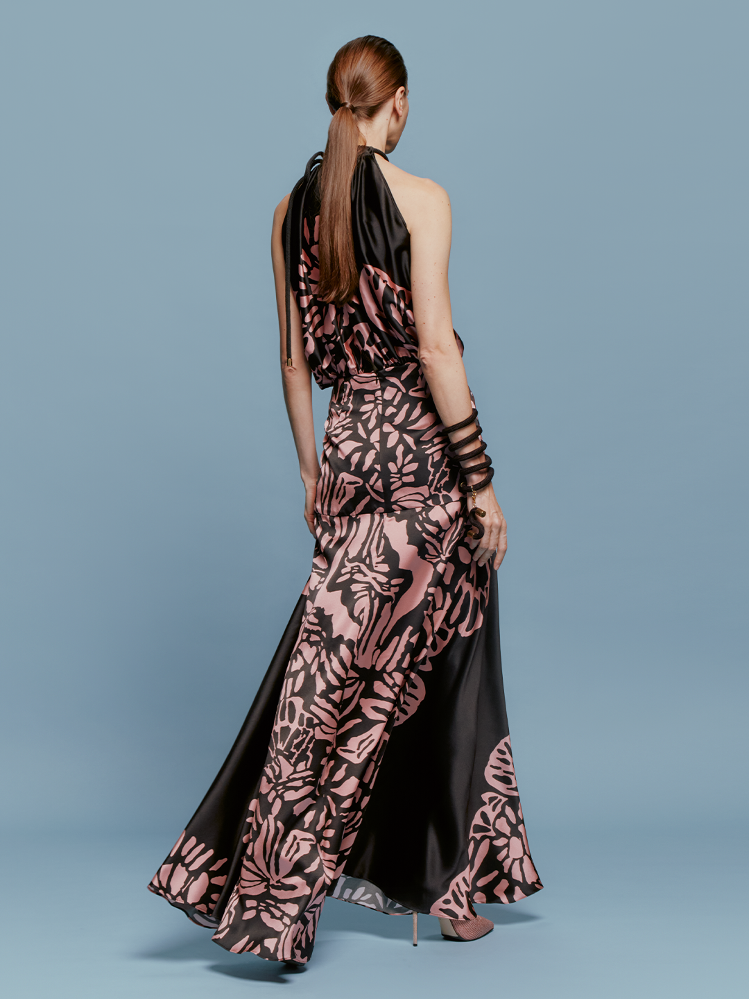 A woman is posing against a blue background, wearing the Joss Dress in Black Pink Filigrana—a black and pink floral halter neck gown—complemented by matching pink heels and black bracelets, capturing the fashion essence of September 2.