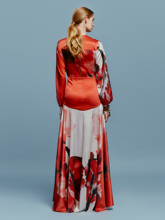 A person is wearing the Keira Dress in Pomegranate Abstract Flowers, featuring long sleeves and a floral print, standing against a plain blue background as if ready for the upcoming events on August 19th.