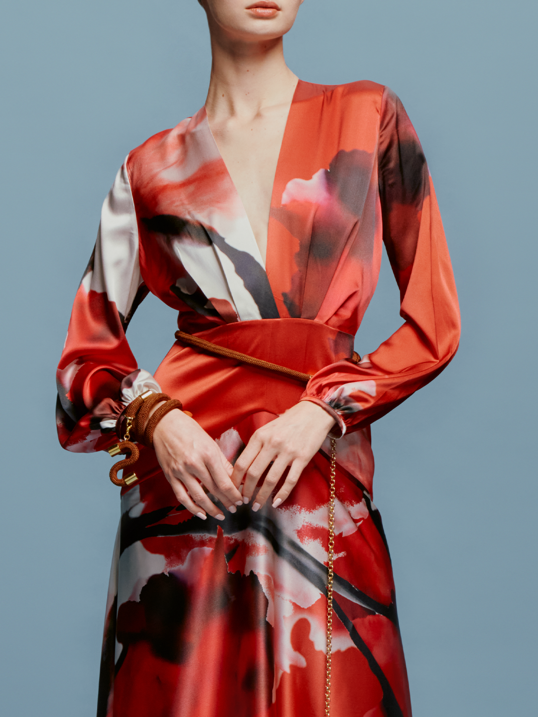 A person is wearing the Keira Dress in Pomegranate Abstract Flowers, featuring long sleeves and a floral print, standing against a plain blue background as if ready for the upcoming events on August 19th.