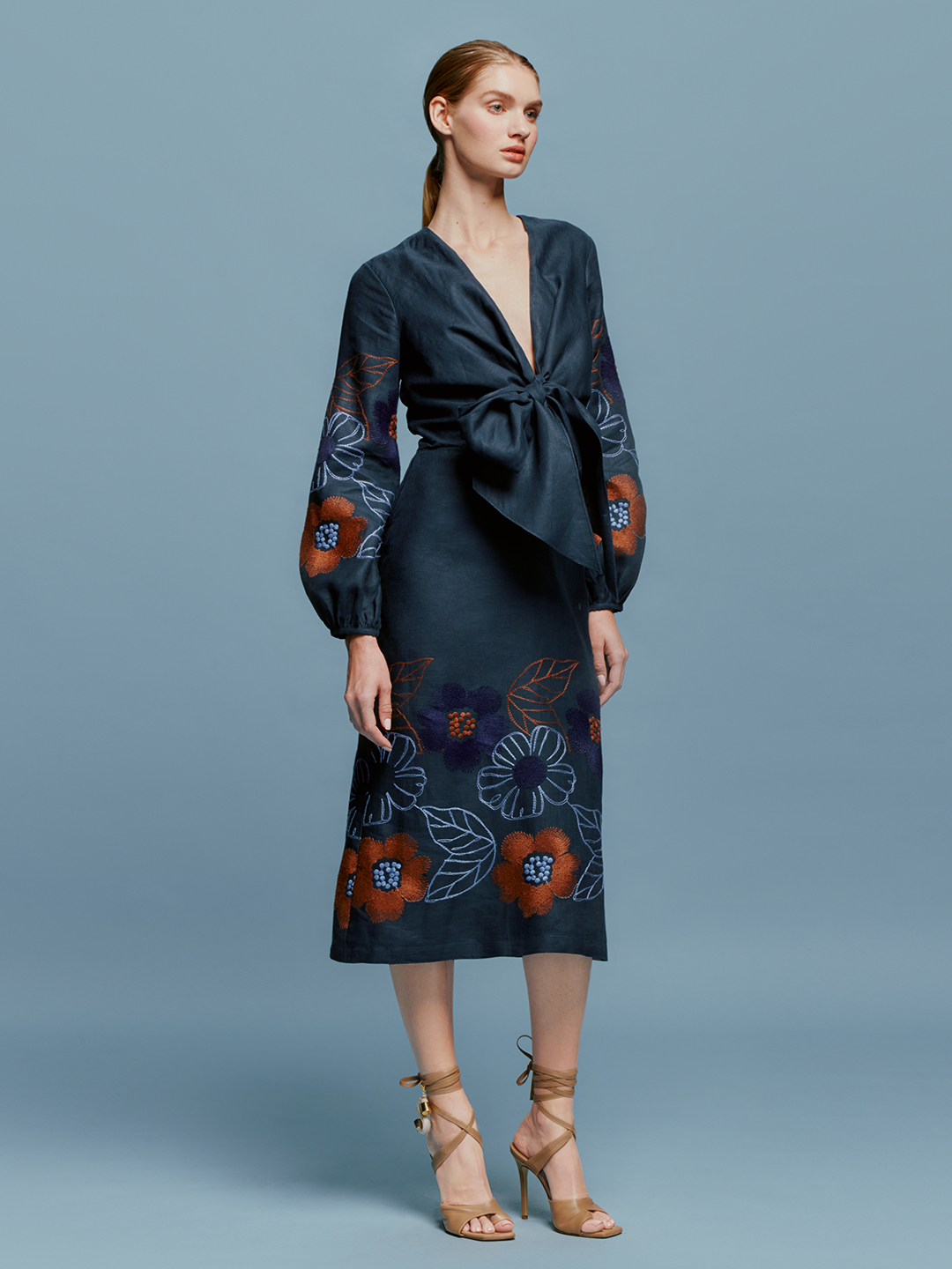 A person stands against a blue background wearing the Laoise Dress in Navy Ochre Flower Embroidery, paired with beige high-heeled sandals, perfect for an August 19th or September 2nd outing.